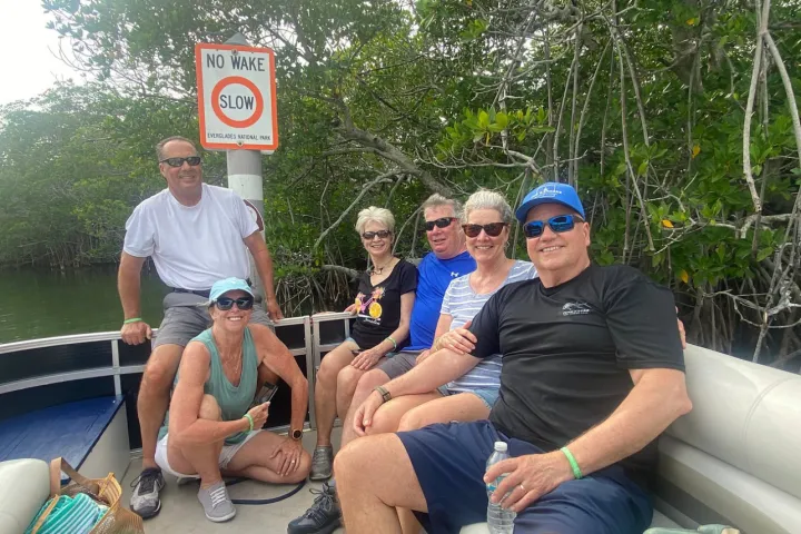 Group of six people on a boat near mangroves with a 'No Wake Slow' sign.