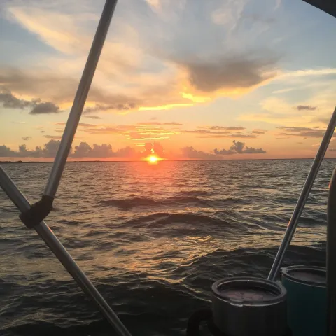 Sunset over ocean with orange sky, viewed from a boat's deck with railing and drinks in foreground.